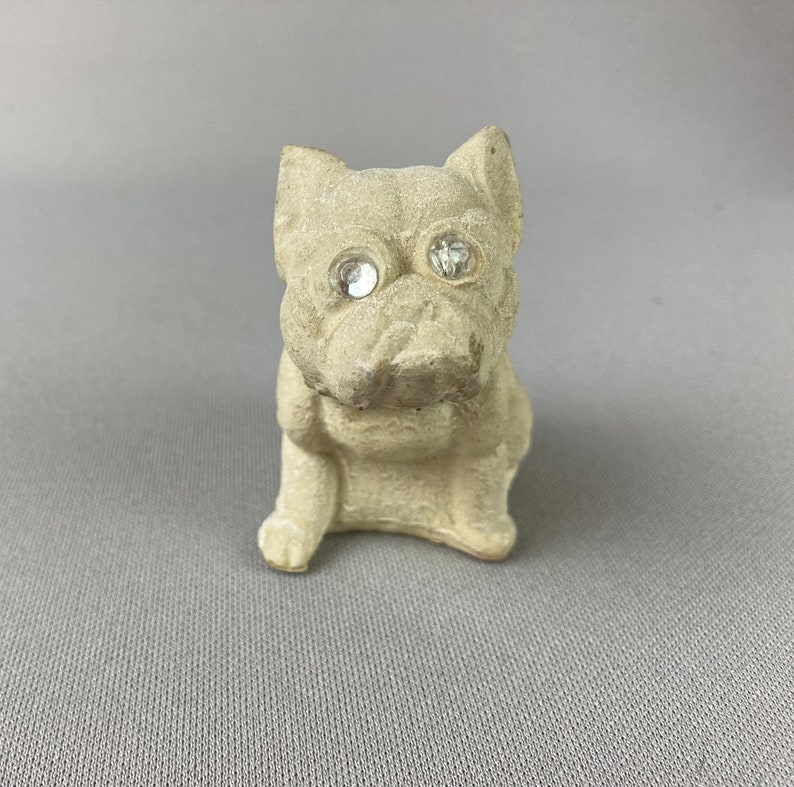 Pu&ograve; includere: Una piccola statuetta di ceramica bianca di un cane seduto con la testa leggermente inclinata di lato. Il cane ha due occhi di strass trasparenti.