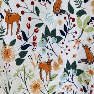 May include: White fabric with a repeating pattern of orange foxes and deer, along with flowers and foliage in shades of green, blue, and orange. The design includes berries and leaves, creating a whimsical forest theme.