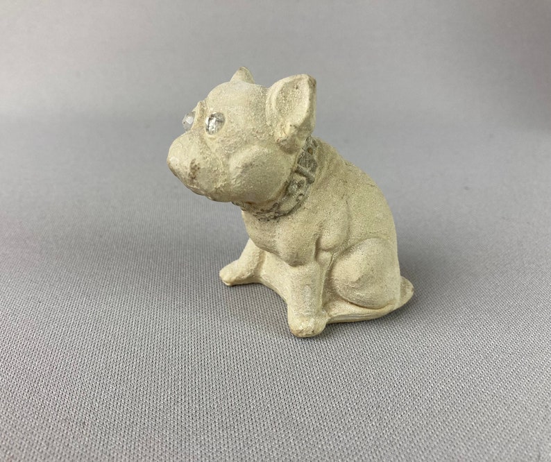 Pu&ograve; includere: Una piccola statuetta in ceramica bianca di un bulldog francese con un collare gioiello. La statuetta ha un aspetto realistico e occhi di strass scintillanti.