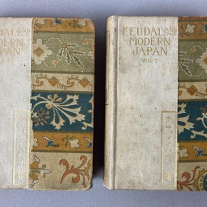 May include: Two antique books titled "Feudal and Modern Japan," Volume 1 and Volume 2. The book covers are off-white with decorative patterned borders in shades of green, blue, and gold. Japanese text is visible on the spine.