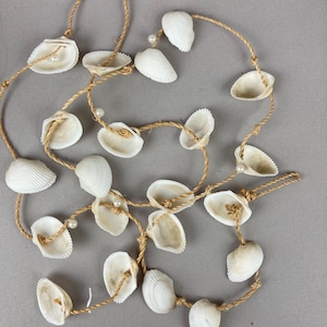 May include: A decorative garland featuring white seashells strung along a natural tan jute rope. The arrangement includes small, round, white faux pearls interspersed between the shells, which are attached to the cord at regular intervals.
