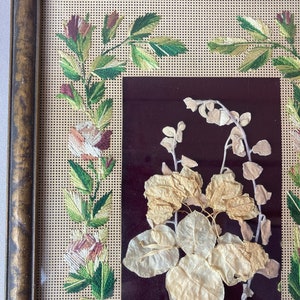 Op de afbeelding: Een ingelijst borduurwerk met een bloemenmotief. De borduurwerk is uitgevoerd in tinten groen, wit en roze. De lijst is van hout en heeft een donkerbruine afwerking.