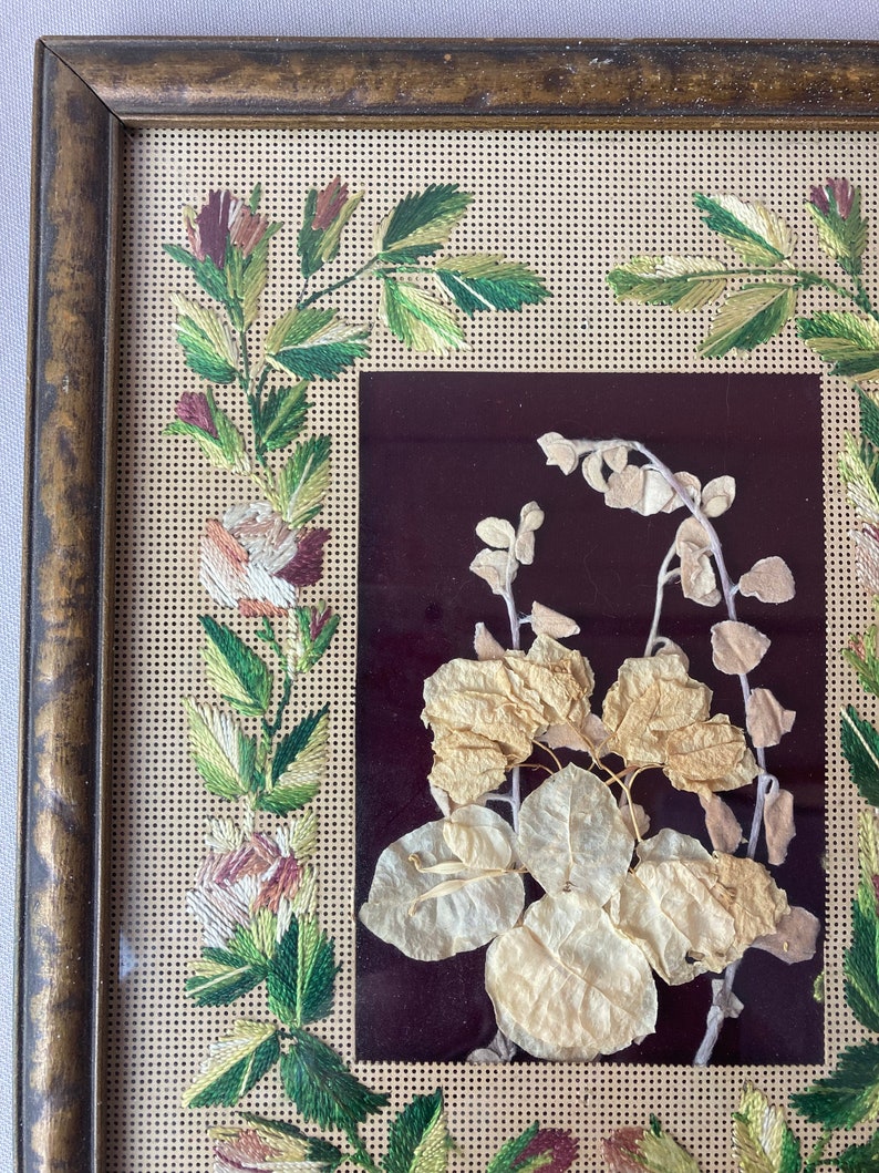 Op de afbeelding: Een ingelijst borduurwerk met een bloemenmotief. De borduurwerk is uitgevoerd in tinten groen, wit en roze. De lijst is van hout en heeft een donkerbruine afwerking.