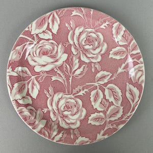 May include: Round, pink and white floral plate. The plate features a repeating pattern of white roses and leaves on a pink background. The plate has a smooth, glossy finish and a white rim. This decorative plate is perfect for serving food or as a decorative piece.