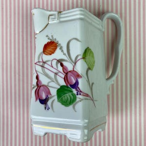 May include: White porcelain pitcher with a rectangular shape and a handle. The pitcher is decorated with a floral design featuring pink, purple, orange, and green flowers and leaves. Gold trim accents the base and rim.