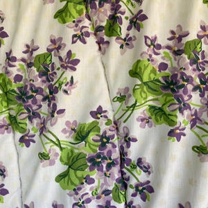 May include: White fabric with a repeating floral pattern of purple violets and green leaves. The design features small, delicate flowers and foliage, creating a vintage aesthetic. The background has a subtle dotted texture.