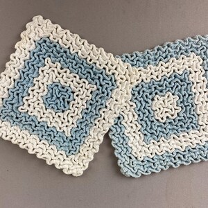 May include: Two square crochet coasters featuring a textured, wavy pattern. Each coaster displays alternating concentric bands of off-white and light blue yarn, arranged in a repeating geometric square design with a ruffled, dimensional surface texture.