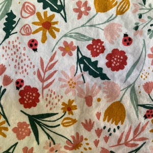 May include: A white fabric with a colorful floral pattern. The design features various flowers in shades of red, pink, and yellow, along with green leaves and small ladybugs. The overall aesthetic is a cheerful, springtime theme.