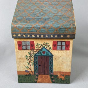 May include: A hand-painted wooden box shaped like a house with a blue roof, yellow walls, red shutters, a brown door, and a vine growing up the side. There is a cat painted on the lawn in front of the house.