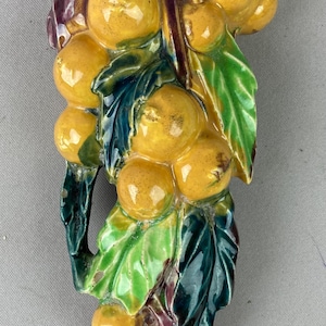 May include: A ceramic wall pocket featuring a cluster of yellow citrus fruits with green leaves and dark purple stems. The glossy, hand-painted piece displays a vertical arrangement of round fruits nestled among textured foliage.