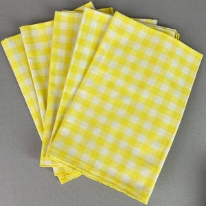 May include: A set of six yellow and white gingham napkins. The napkins are folded and stacked on a gray surface.