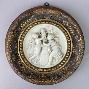 May include: A round, antique wall plaque with a bas-relief scene of three figures and a child. The central relief is white, surrounded by a gold beaded border and a dark brown frame with gold floral accents. A small metal loop is at the top.