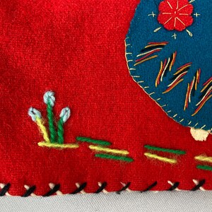 以下が含まれることがあります： 赤色のフェルト生地に、青と黄色の刺繍が施されています。デザインは、花、線が入った円、白い花がついた緑色の茎で構成されています。