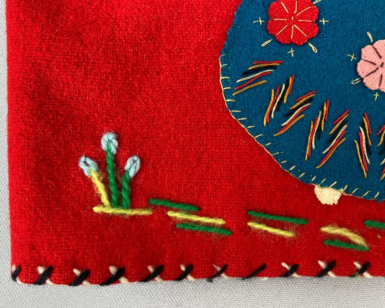 以下が含まれることがあります： 赤色のフェルト生地に、青と黄色の刺繍が施されています。デザインは、花、線が入った円、白い花がついた緑色の茎で構成されています。