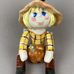May include: A ceramic figurine of a scarecrow wearing a tan hat with a black crow on top, yellow hair, and a yellow plaid shirt. The figure has blue eyes, pink cheeks, and brown pants with black boots, while holding a small orange pumpkin.