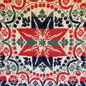 May include: Close-up of a vintage textile with a repeating pattern. The design features a central star shape in red, green, and navy blue, surrounded by floral and geometric motifs in the same colors on a cream background. The textile has a woven texture.