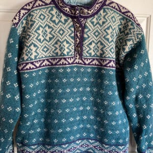 May include: A teal, cream, and purple patterned sweater with a high collar and buttoned placket. The sweater features a geometric design around the collar and chest, with a diamond pattern on the body. The sleeves are long.