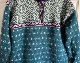 Könnte beinhalten: Ein türkisfarbener, cremefarbener und lila gemusterter Pullover mit hohem Kragen und Knopfleiste. Der Pullover hat ein geometrisches Design um den Kragen und die Brust, mit einem Rautenmuster auf dem Körper. Die Ärmel sind lang.