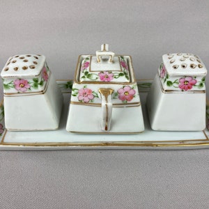 May include: A white porcelain salt and pepper shaker set with a matching sugar dispenser. The set features a floral design with pink flowers and green leaves. The shakers have holes on top for dispensing salt and pepper. The sugar dispenser has a lid with a handle.