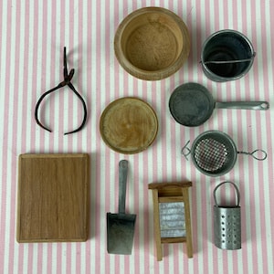 May include: A collection of miniature kitchen tools, including a wooden bowl, a small metal bucket, a frying pan, a strainer, a cutting board, a scoop, a washboard, and a metal container. The items are arranged on a pink and white striped surface.