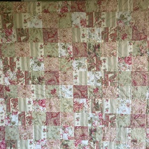 May include: A patchwork quilt featuring a repeating pattern of squares in various floral and paisley prints. The color palette includes shades of red, green, cream, and tan, creating a vintage aesthetic. The quilt is likely handmade.