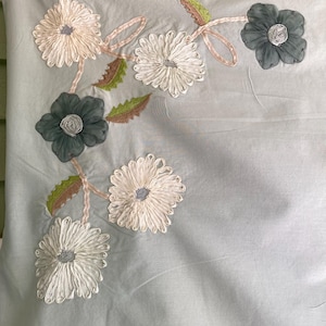 May include: A light gray fabric square featuring decorative floral embroidery. The design includes white and dark gray fabric flowers with textured petals, connected by light pink ribbon stems and accented with small green serrated leaves.