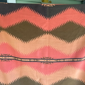 May include: A rectangular blanket featuring a repeating geometric pattern of jagged, zigzagging stripes in shades of dark brown, muted orange, and coral pink. A horizontal band with intricate, symmetrical dark brown motifs runs across the center.