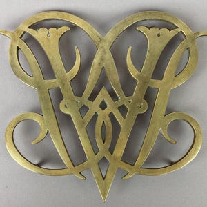 May include: A brass trivet with an ornate, openwork design. The trivet features a stylized, intertwined "W" and "V" monogram, with floral accents and curved flourishes. The metal has a warm, golden hue and shows signs of age.
