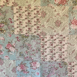 May include: A quilted fabric blanket featuring a patchwork design of various floral patterns in shades of dusty rose, sage green, and cream. The textile displays repeating motifs of roses, small blossoms, and vines across square panels with visible stitching.