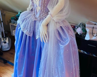 May include: A lavender and blue ball gown displayed on a mannequin. The bodice is embellished with white embroidery, pearls, and pink floral accents. The skirt features layers of satin and tulle, with a delicate pearl overlay. A matching choker and armbands complete the ensemble.