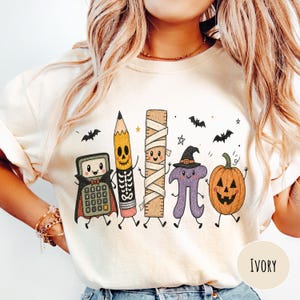 May include: An ivory t-shirt featuring a graphic print of five animated school supplies for Halloween. The characters include a calculator in a vampire cape, a skeleton pencil, a mummy ruler, a pi symbol in a witch hat, and a jack-o'-lantern.