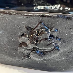 Pode incluir: Uma tigela de cerâmica cinza-carvão escuro com uma escultura em relevo brilhante de um rosto de gato na parede externa. A tigela tem um acabamento fosco e texturizado na superfície externa, enquanto o detalhe do gato é liso, brilhante e reflexivo.