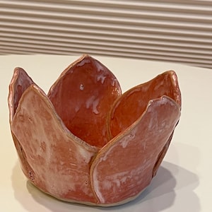 Puede incluir: Un cuenco de cerámica con forma de flor abierta y seis pétalos curvos distintos. Las superficies exterior e interior presentan un esmalte brillante de color rosa coral moteado con sutiles reflejos blancos en los bordes y texturas irregulares.