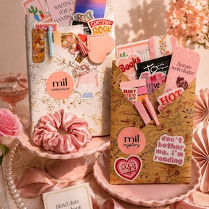 May include: Two paper envelopes filled with book-themed stationery, stickers, and bookmarks. The white envelope features gold floral patterns and a pink scrunchie. The brown envelope has a botanical print. A card reads, blind date with a book.