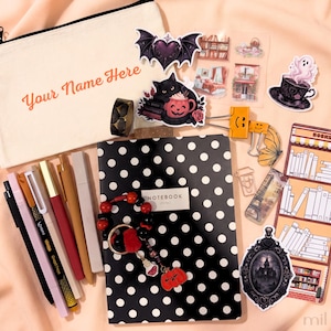 May include: A stationery set including a cream-coloured pencil case with the text Your Name Here, a black and white polka dot notebook, various pens, a beaded keychain with a pumpkin charm, and assorted stickers featuring bats, ghosts, and books.