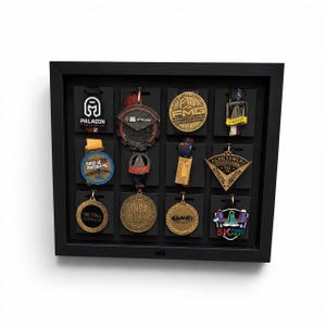 May include: A black rectangular shadow box display case containing twelve assorted sports and fitness medals. The medals are arranged in a grid on black backing cards, featuring various shapes, metallic finishes, and colourful ribbons.