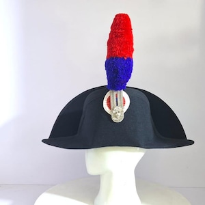 Op de afbeelding: Een zwarte steekhoed met een opvallende rode en blauwe pluim aan de voorzijde. De pluim is bevestigd met een zilverkleurig metalen embleem in het midden van een ronde wit-rode kokarde, wat zorgt voor een formele en kenmerkende uitstraling.