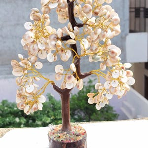 May include: A decorative tree sculpture featuring a dark brown trunk and branches made of thin gold-colored wire. The branches are adorned with numerous small, round, off-white and tan seashells. The base is a cylindrical wooden block.