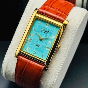May include: A rectangular Seiko Quartz watch with a gold-tone case and a bright turquoise dial featuring a wavy texture pattern. The dial displays gold-tone Roman numeral hour markers and hands, paired with a textured orange leather strap.