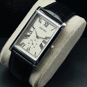 May include: A rectangular Seiko wristwatch featuring a silver-toned case, a white dial with black Roman numeral hour markers, and a black leather strap with a textured finish. The watch includes a small sub-dial for seconds and silver hands.
