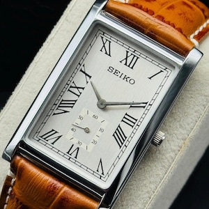 Peut inclure: Une montre-bracelet rectangulaire Seiko avec un cadran argenté, des chiffres romains noirs et un sous-cadran pour les secondes. La montre présente un boîtier en métal argenté poli et un bracelet en cuir fauve texturé avec des coutures blanches.
