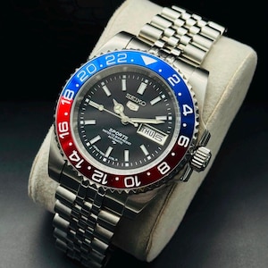 May include: A stainless steel Seiko 5 Sports automatic wristwatch featuring a black dial with luminous markers, a day-date window at the three o'clock position, and a rotating bezel with a red and blue insert. It has a silver-tone jubilee bracelet.