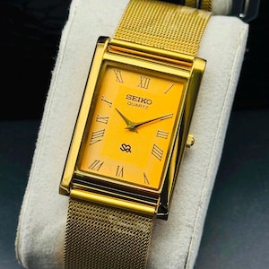 May include: A rectangular gold-tone wristwatch featuring a vibrant yellow dial with black Roman numeral hour markers. The watch has a textured gold-tone mesh strap and displays the text SEIKO QUARTZ on the face with a small logo below the center.