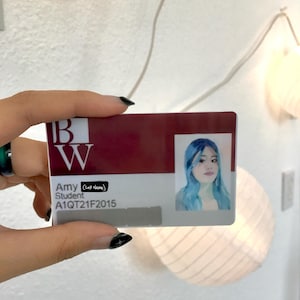 May include: A rectangular student identification card featuring a maroon header with the letters BW in white. The card displays the name Amy, the title Student, the ID number A1QT21F2015, and a portrait of a person with blue hair on the right side.
