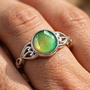 May include: A silver-toned ring featuring a round, color-changing mood stone set in a bezel. The band is crafted with an openwork, swirling filigree design that extends from the central setting, creating a detailed and intricate metal structure.