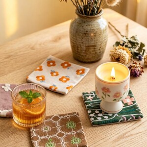 Könnte beinhalten: Eine Sammlung quadratischer Stoffuntersetzer mit verschiedenen Stickmustern, darunter orangefarbene Blumen auf Creme, weiße geometrische Muster auf Braun und rosa Blumen auf Dunkelgrün, neben einem Glas Orangensaft und einer Kerze.
