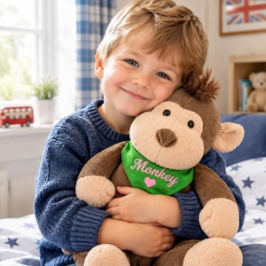 May include: A plush brown monkey stuffed animal with a tan face and ears, wearing a bright green triangular bandana around its neck. The bandana features the embroidered word Monkey in pink script above a small pink heart icon.