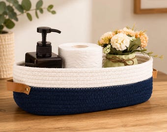 Op de afbeelding: Een ovale opbergmand van gewikkeld touw met een wit bovengedeelte en een donker marineblauw ondergedeelte. De mand is voorzien van beige kunstlederen handvatten aan beide zijden, bevestigd met goudkleurige metalen klinknagels.