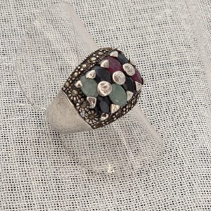 May include: A silver ring featuring a cluster of oval-shaped gemstones in shades of red, black, and pale green. The setting is accented with small, dark, metallic circular studs arranged between the stones, creating a textured, multi-faceted appearance.