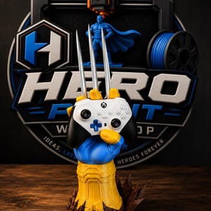 May include: A blue and yellow sculpted hand stand holding a white and black game controller with three silver metallic claws extending upward. The base features a circular logo with the text HERO PRINT SHOP, IDEAS. LAYERED. HEROES FOREVER.